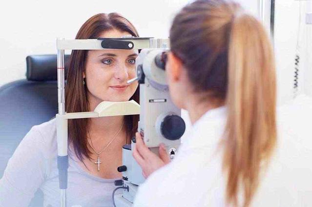 The camera focuses on a young woman looking through a retinal camera as her optician looks through the other side
